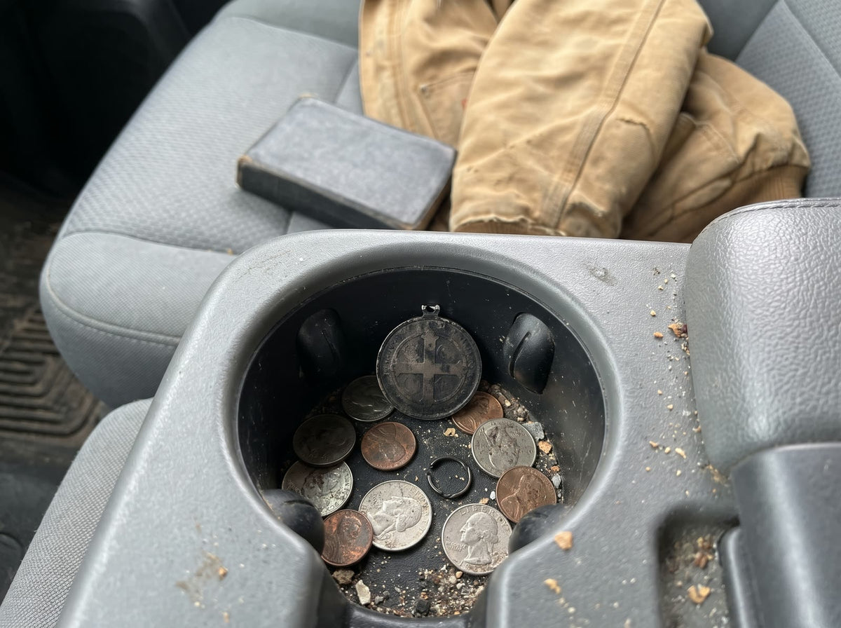 A cheap black medal with its ring snapped off among coins in a pickup’s cup holder, a prayer book on the seat