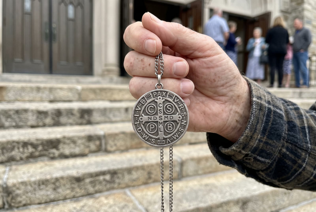 An older man’s hand holding up a worn St Benedict medal on the church steps
