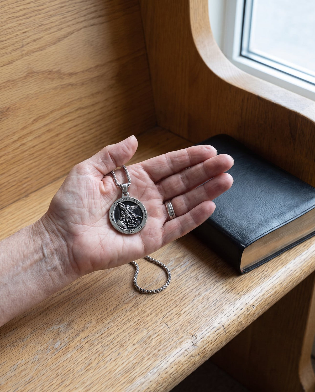 The silver St Michael medal in a woman’s open palm over a pale oak pew beside a closed missal