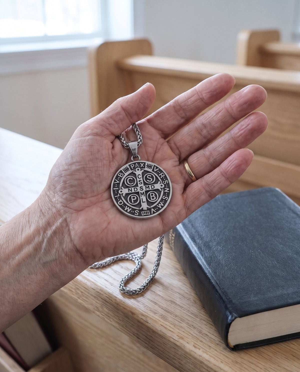 The St Benedict medal in a woman’s palm over a pale oak pew, a missal beside it