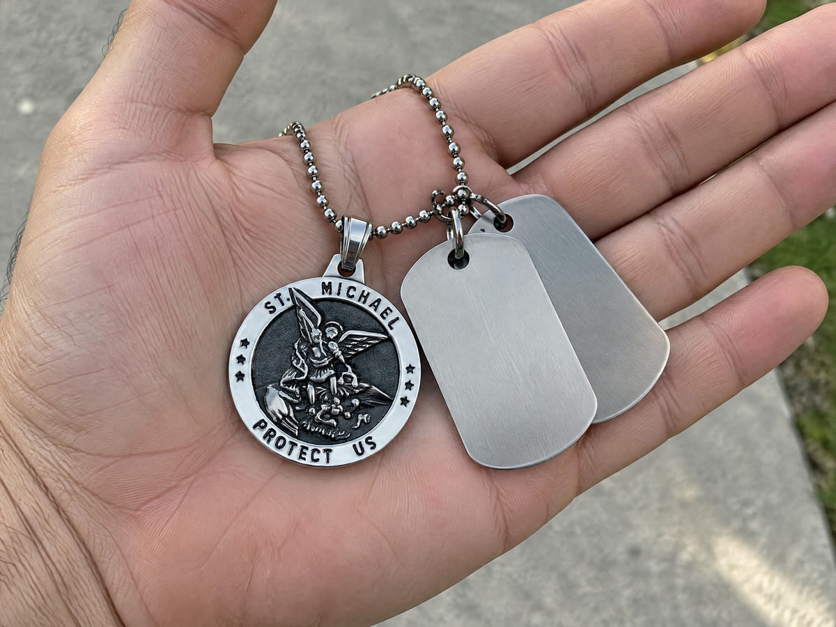A man’s open palm holding a silver St Michael medal on a ball chain beside two blank dog tags