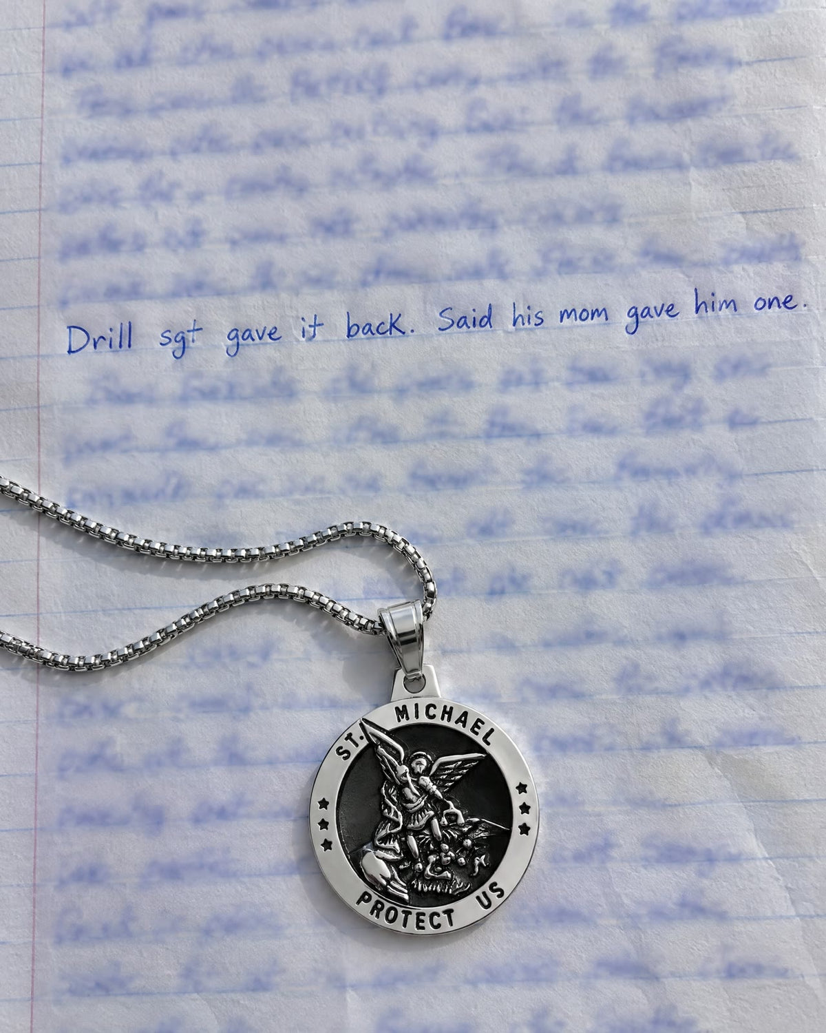 A handwritten letter on notebook paper reading Drill sgt gave it back. Said his mom gave him one., a silver St Michael medal on the page