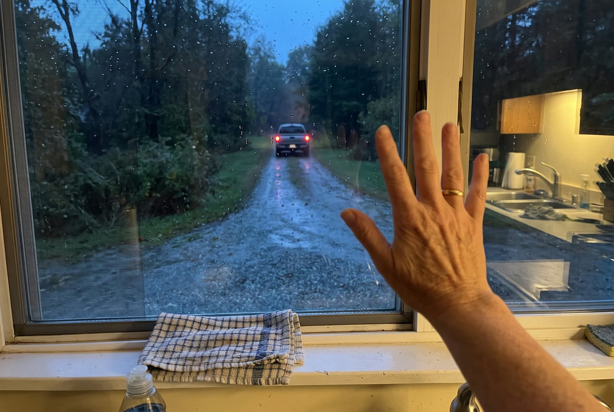 A woman’s hand on a rainy kitchen window, a pickup’s taillights at the end of the driveway