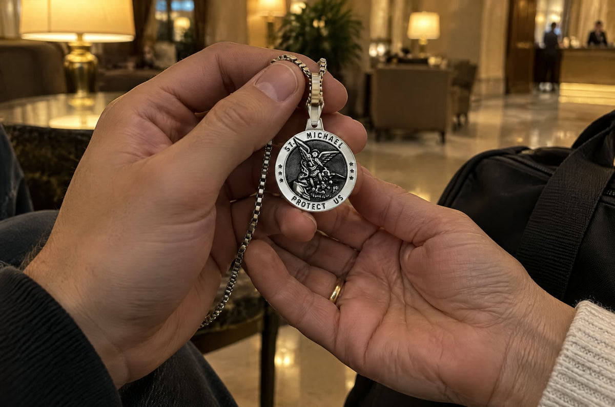 In a hotel lobby at night, a young man’s hand turns a silver St Michael medal to read the rim, his mother’s hand cupped under his