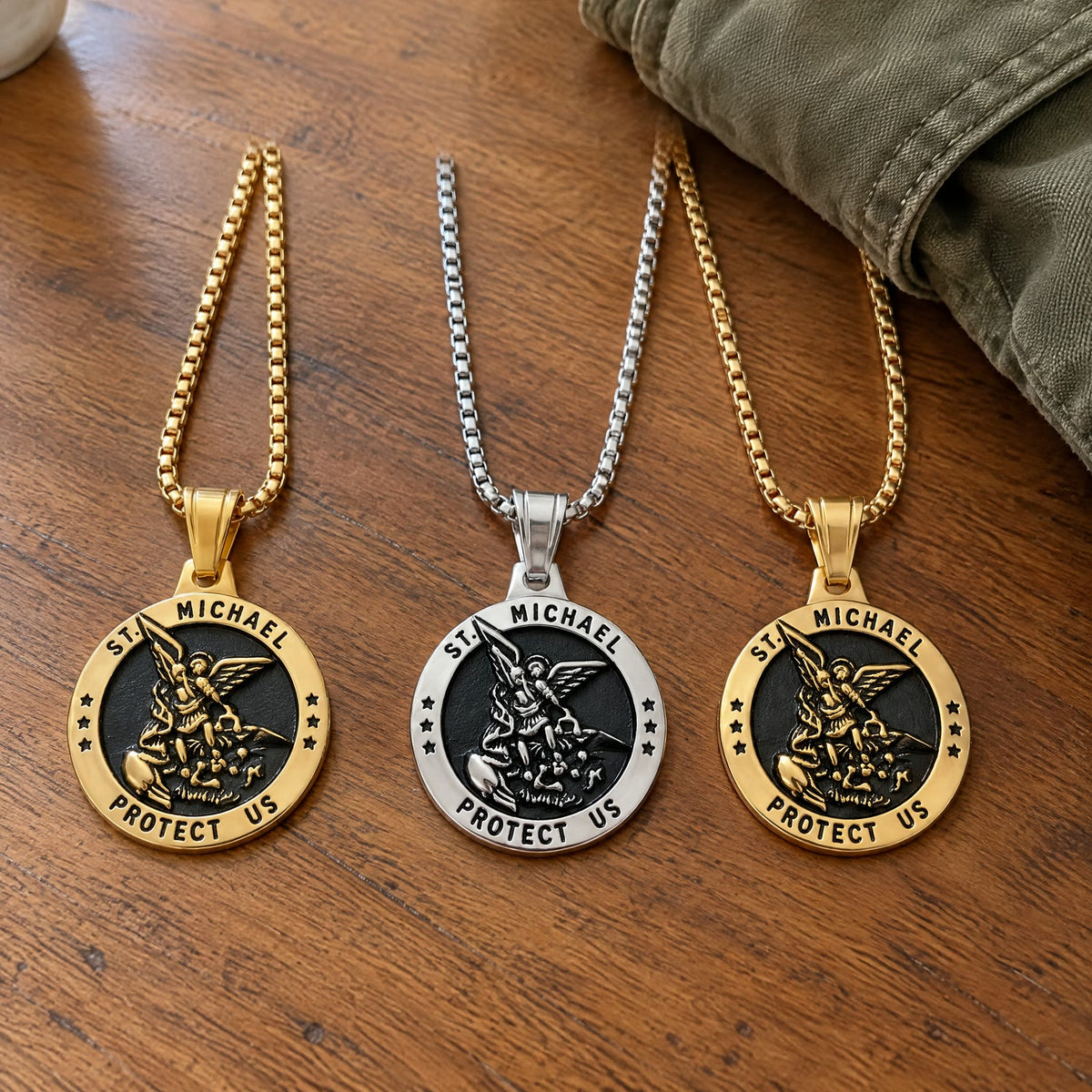 Three identical St Michael medals on box chains, two gold and one silver, on a wooden kitchen table beside a folded jacket