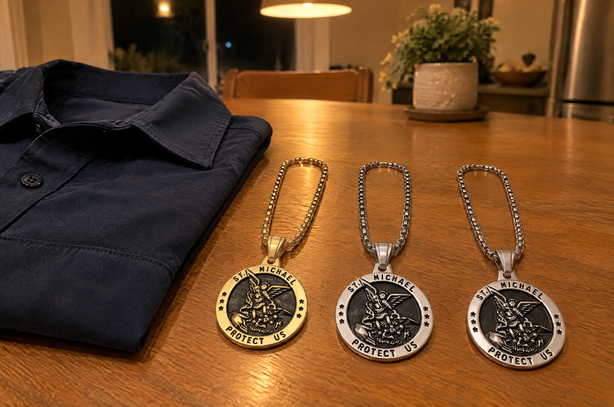 Three St Michael medals, one gold and two silver, on a kitchen table at night beside a folded navy uniform shirt